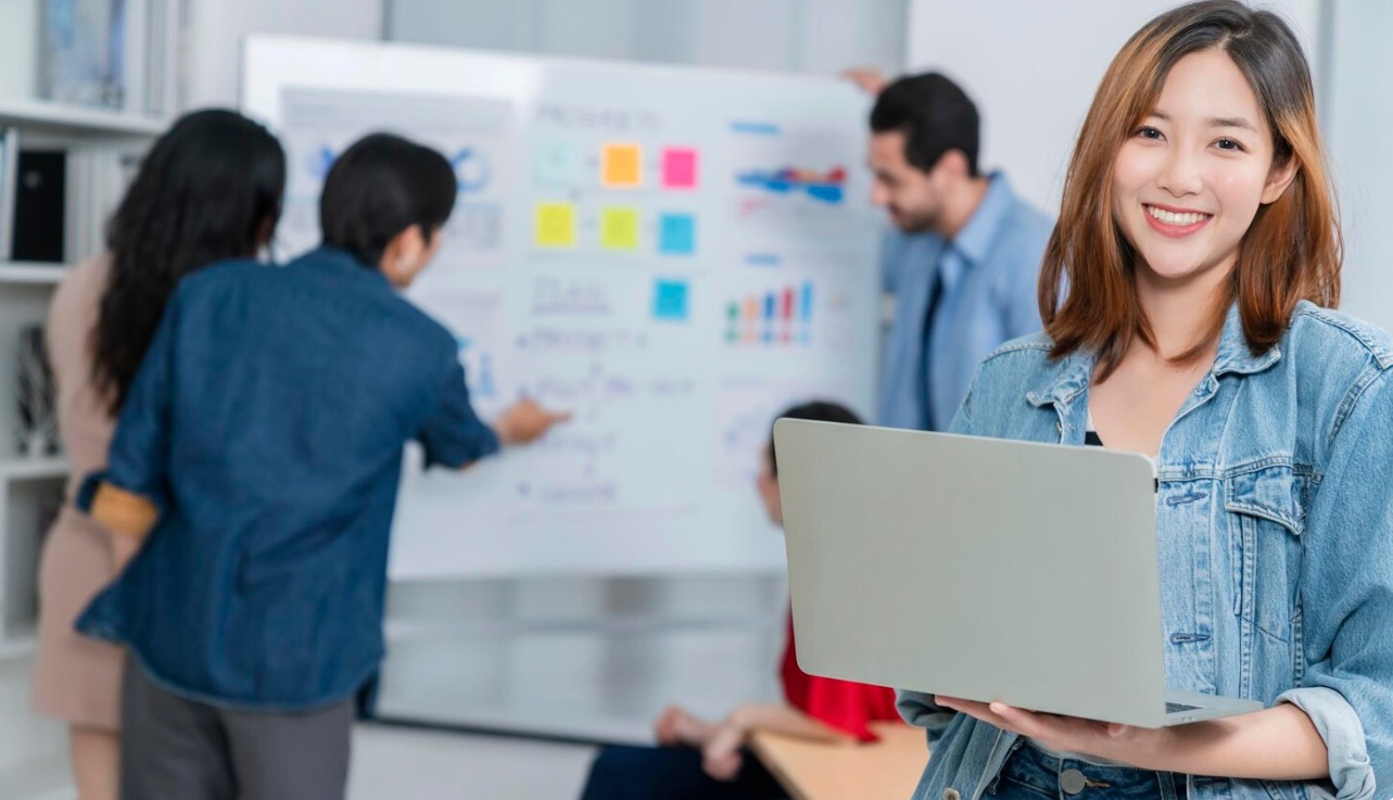 Asian entrepreneur standing with her laptop in front of team mates working on a white board
