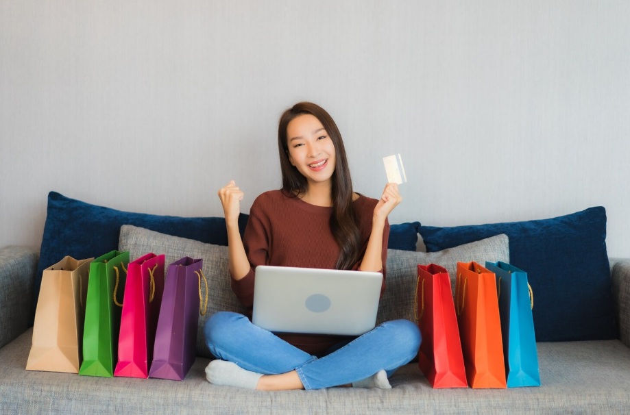 Young Asian woman sitting on a sofa with her laptop and credit card while surrounded by colored paper bags
