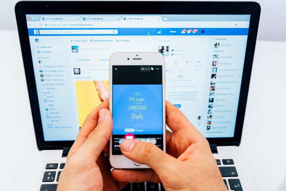 Man holding phone in front of a monitor displaying Facebook pages